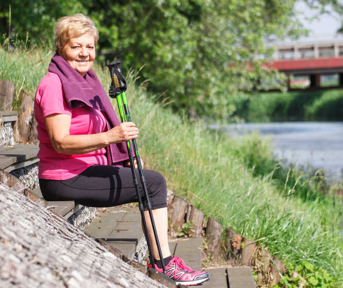 Bei der Herzschwäche-Behandlung ist auch Bewegung wichtig. Eine Patientin mit Herzschwäche macht Rast in der Natur und hält zwei Nordic-Walking-Stöcke in der Hand.